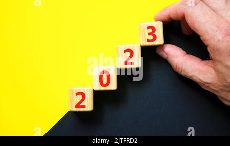 2023 felice anno nuovo simbolo. I cubi di legno simboleggiano il passaggio dal 2022 al nuovo anno 2023. Mano uomo d'affari. Splendido sfondo giallo e nero. Foto Stock