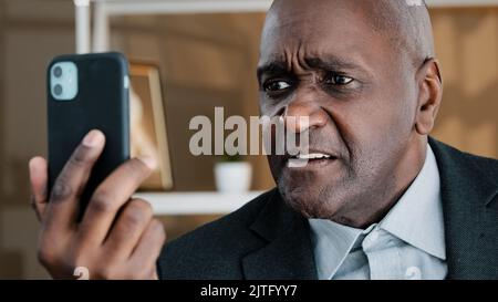 Arrabbiato anziano uomo d'affari africano che ha emotivamente spiacevole conversazione videochiamata con smartphone sconvolto fallimento boss dipendenti cattive notizie Foto Stock