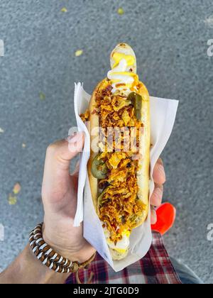 L'uomo tiene in mano un cane caldo con cipolle fritte e peperoni. Messa a fuoco selettiva. Cibo di strada. Foto verticale Foto Stock