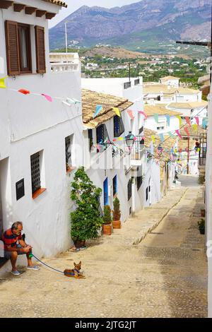 Un uomo e il suo cane si siedono su una collina fiancheggiata da case dalle pareti bianche drappeggiate da bandiere colorate ad Altea, Alicante, Spagna Foto Stock