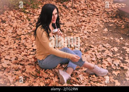 Donna brunetta in t-shirt gialla jeans blu snd seduta su foglie arancioni in autunno Foto Stock