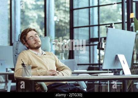 Stanco TECNICO specialista IT programmatore dorme in ufficio al tavolo durante il giorno, uomo in occhiali e abiti casual lavora al computer, uomo d'affari riposa Foto Stock