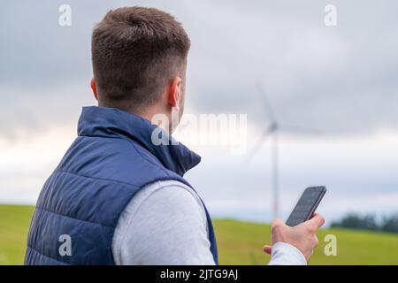 Il tecnico utilizza il telefono cellulare per controllare il funzionamento della turbina eolica. La turbina eolica produce energia verde contro il cielo grigio Foto Stock