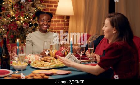 Felice diverse donne che si godono la cena di Natale insieme mentre chiacchierano. Gente multietnica gioiosa che festeggia le vacanze invernali con il cibo tradizionale casalingo cucinato mentre si sente bene. Foto Stock