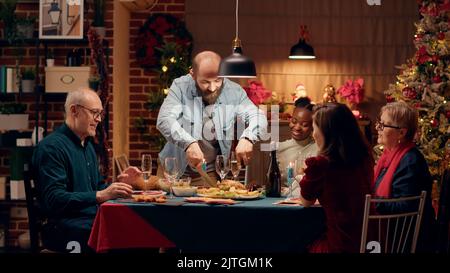 Fiero marito tagliare pollo arrosto per gli ospiti mentre godersi la cena di Natale a casa. Festive uomo che si gode la vacanza invernale con tradizionale cibo casalingo e champagne. Foto Stock