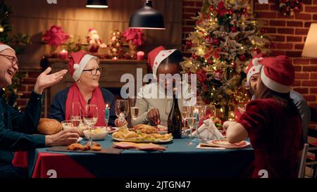 Persone diverse e positive sorridono di cuore mentre si gustano la cena di Natale a casa con i propri cari. I membri della famiglia festeggiano la festa invernale con piatti tradizionali fatti in casa. Scatto manuale Foto Stock