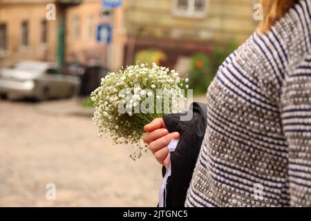 Defocus femmina mano che tiene mazzo di fiori bianchi. Primo piano di piccoli fiori bianchi di gypsofila. Twig con fiori di Gypsophila sullo sfondo della città. Fuori Foto Stock