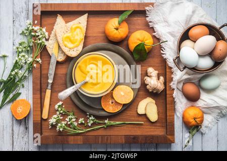 Vasetto di cagliata d'arancia su vassoio di legno con pane, arance, zenzero e uova in un recipiente di rame Foto Stock