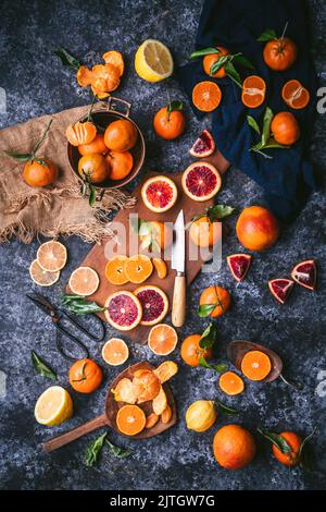 Piatto di vari agrumi colorati disposti su uno sfondo scuro con tagliere rustico e coltello Foto Stock