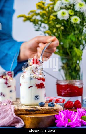 Donna che prende un cucchiaio di yogurt da un piccolo vasetto riempito con yogurt prima colazione parfait con confettura di fragole, farinata d'avena, lamponi, mirtilli. Foto Stock
