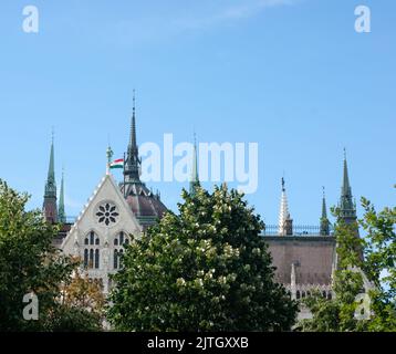 Parlamento a Budapest, Ungheria, siede con guglie, torri, tetto a picco, E la bandiera ungherese. Sfondo cielo blu con alberi in primo piano. Foto Stock