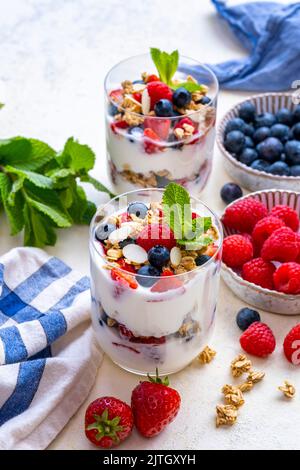 Yogurt e parfait di frutta serviti in due bicchieri e accompagnati da fragole, mirtilli, lamponi, muesli e foglie di menta fresca. Foto Stock