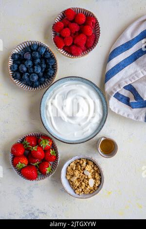 Yogurt greco, lamponi, mirtilli, fragole, muesli e sciroppo d'acero fotografati in ciotole di ceramica su uno sfondo chiaro. Foto Stock