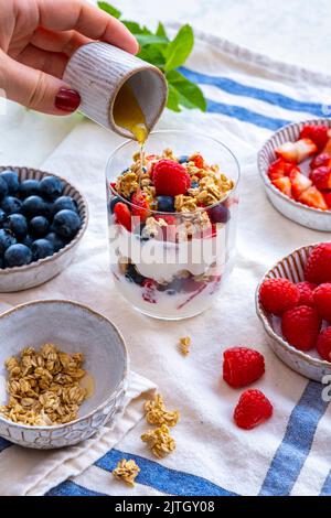 Donna mani versare sciroppo d'acero su yogurt parfait in un bicchiere, mirtilli, lamponi, fragole e farinata d'avena in ciotole di ceramica sul lato. Foto Stock