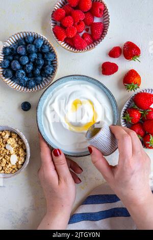 Donna mani versare sciroppo d'acero su yogurt greco, lamponi, fragole, mirtilli e muesli sul lato. Foto Stock