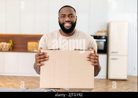 Felice giovane afro-americano, in piedi a casa nel salotto, tenendo una grande scatola di cartone, ricevuto pacco dal negozio online, preparandosi a disimballare, guarda la macchina fotografica, sorriso, eccitazione Foto Stock