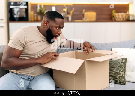 Scioccato confuso africano americano ragazzo di apertura grande scatola di cartone, disimballaggio internet ordine negozio a casa, si sente frustrato con il lungo servizio di consegna e le merci che hanno ricevuto, esperienza di sconcertante Foto Stock