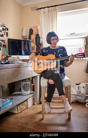 È il momento di sdraiarsi dei brani. Una giovane donna elegante seduta nella sua eclettica sala da gioco suonando una chitarra. Foto Stock