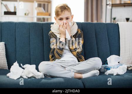 Concetto di coronavirus, polmonite e influenza. Vista frontale di un ragazzino malato esausto, seduto sul divano di casa, soffia il naso che corre nel fazzoletto di carta, ha difficoltà a respirare, si sente male. Foto Stock