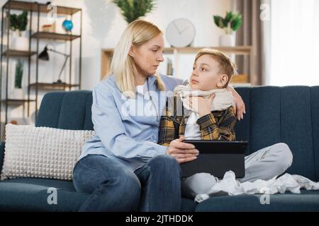 Concetto di supporto familiare durante la malattia. Piacevole madre di mezza età che passa il tempo a casa insieme al suo figlio adolescente malato, seduto sul divano e usin Foto Stock