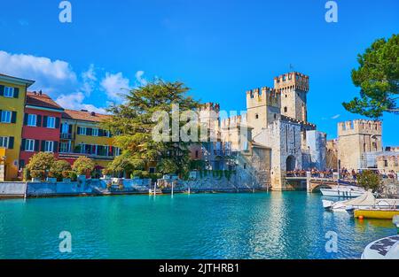 Il porticciolo della località turistica di Sirmione con vista su belle case colorate, alberghi, Castello Scaligero e piccole barche, ormeggiate in riva al fiume, Lomba Foto Stock