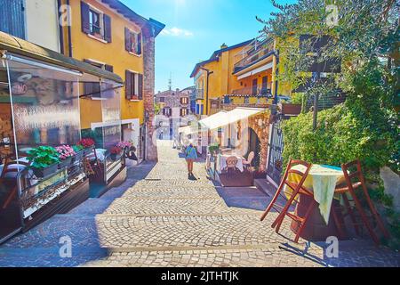 Passeggia lungo la stretta Via San Salvatore con caffè all'aperto, ristoranti e bar, decorati con fiori in vaso e piante verdi, Sirmione, Ita Foto Stock