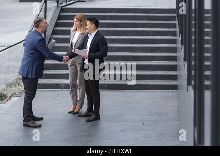 Incontro di tre colleghi esterni all'edificio dell'ufficio, specialisti IT esperti e maturi, accoglienza e scuotimento delle mani, persone aziendali in tute aziendali, gruppi diversi di persone Foto Stock