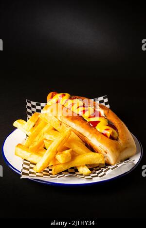 Salsiccia di Francoforte con ketchup e senape accompagnata da patatine fritte su un piatto bianco su sfondo nero Foto Stock