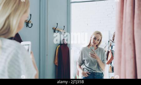 Bella donna bionda sta facendo selfie specchio con telefono intelligente mentre si trova in piedi in una bella stanza vestito in boutique di lusso vestiti. Sta posando, muovendosi e sorridendo con noncuranza. Foto Stock