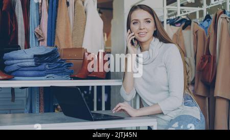 Giovane donna d'affari che lavora con il portatile e che parla allegramente sul suo telefono cellulare seduto nel negozio di abbigliamento. Abbigliamento elegante in esposizione e il cliente si muove sullo sfondo. Concetto di piccola impresa. Foto Stock