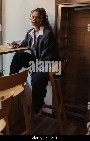 elegante donna afroamericana in tuta di grandi dimensioni che tiene uno smartphone mentre si siede in un bar Foto Stock