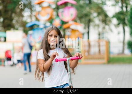 Una bella ragazza sorridente percorre uno scooter lungo il sentiero di un parco divertimenti in una giornata estiva. Sport stagionale per bambini. Foto Stock