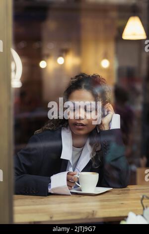 donna afroamericana riccia che tiene una tazza di caffè dietro il vetro della finestra nel caffè Foto Stock