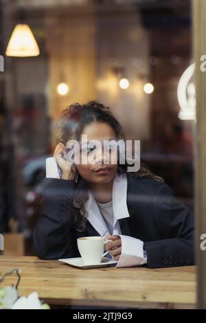 donna afro-americana penosa che tiene una tazza di caffè dietro il vetro della finestra nel caffè Foto Stock