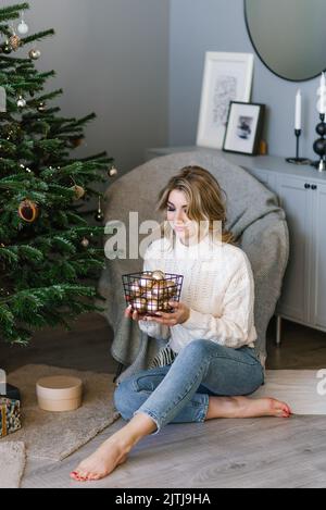 Bella giovane donna in un maglione accogliente e jeans decorare un albero di Natale con palloncini da un cesto a casa Foto Stock