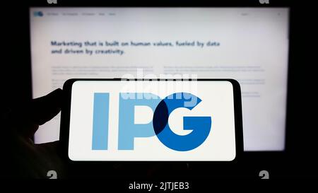 Persona che tiene uno smartphone con il logo di Interpublic Group of Companies Inc. (IPG) sullo schermo di fronte al sito Web. Messa a fuoco sul display del telefono. Foto Stock