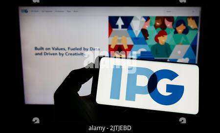 Persona che tiene il cellulare con il logo di Interpublic Group of Companies Inc. (IPG) sullo schermo di fronte alla pagina Web aziendale. Messa a fuoco sul display del telefono. Foto Stock
