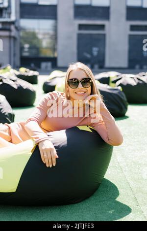 La giovane donna è seduta su una poltrona all'aperto e si gode una giornata di sole. Una bella ragazza contenta in occhiali da sole. Foto Stock