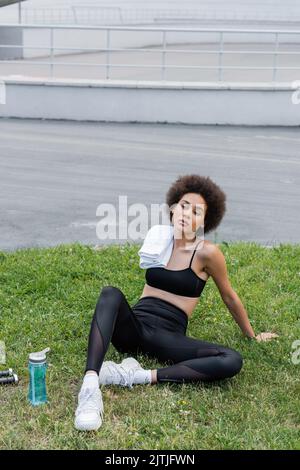 sport afroamericano con asciugamano seduto sul prato vicino a bottiglie sportive e manubri Foto Stock