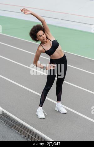 atletica afroamericana donna in abbigliamento sportivo nero facendo l'esercizio di curvatura laterale con mano su anca e mano sollevata Foto Stock