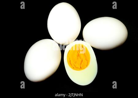 Uova bianche, una delle quali bollita e spezzata a metà su fondo nero Foto Stock