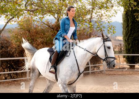 vista laterale di una ragazza che cavalca un cavallo bianco in stalla in estate Foto Stock