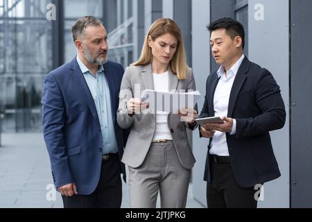 Team aziendale diversificato tre colleghi dipendenti discutono di documenti contratti al di fuori dell'ufficio edificio uomo d'affari e donna d'affari in possesso di fatture e rapporti finanziari parlare Foto Stock