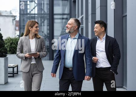 Un team diversificato di specialisti IT, responsabili di ingegneri senior ed esperti, un gruppo di tre lavoratori che si girano felicemente fuori da un edificio di uffici, colleghi in tute aziendali. Foto Stock