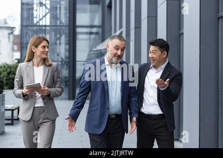 Un team diversificato di specialisti IT, responsabili di ingegneri senior ed esperti, un gruppo di tre lavoratori che si girano felicemente fuori da un edificio di uffici, colleghi in tute aziendali. Foto Stock