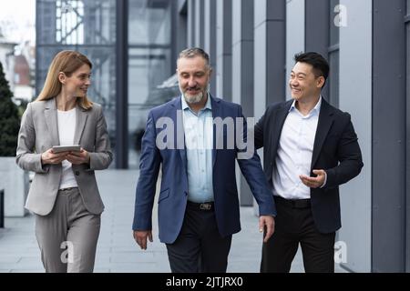 Un team diversificato di specialisti IT, responsabili di ingegneri senior ed esperti, un gruppo di tre lavoratori che si girano felicemente fuori da un edificio di uffici, colleghi in tute aziendali. Foto Stock