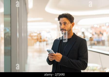 Ritratto di buon uomo d'affari ispanico felice usando il telefono vicino alla finestra al centro commerciale orizzontale sparato Foto Stock