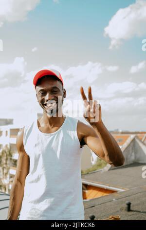 Ritratto di giovane uomo con cappuccio che gestura segno di pace contro il cielo nelle giornate di sole Foto Stock