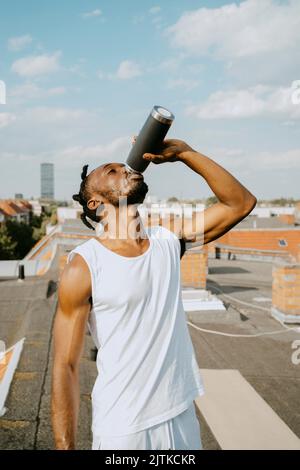 Giovane uomo che beve acqua dalla bottiglia sul tetto durante la giornata di sole Foto Stock