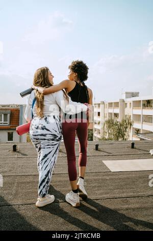Vista posteriore delle amiche che camminano con le braccia sul tetto durante le giornate di sole Foto Stock
